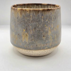 May include: A handmade ceramic tumbler with a cylindrical shape. The upper portion is a mottled gray with streaks of gold and brown, while the base is a speckled beige. The rim is a darker brown.