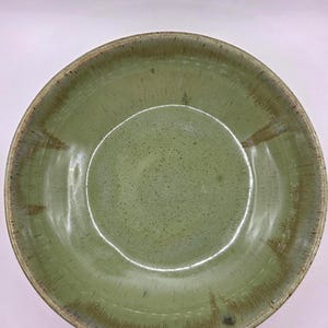 May include: A handmade, round ceramic bowl with a glossy, speckled green glaze. The rim has a darker, earthy brown tone. The bowl's interior is a lighter shade of green, creating a natural, rustic aesthetic. Ideal for serving salads or soups.
