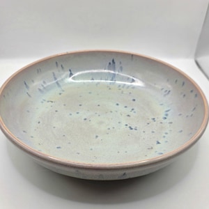 May include: A ceramic bowl with a light blue and cream speckled glaze. The rim is a soft pink, and the bowl has a shallow, rounded shape. This handmade bowl is suitable for serving food.