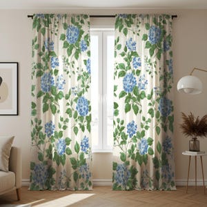 Könnte beinhalten: Blumengardinen mit cremefarbenem Hintergrund, verziert mit blauen Hortensienblüten und grünen Blättern. Die Vorhänge hängen vor einem Fenster und verleihen der Raumdekoration ein natürliches, botanisches Thema.