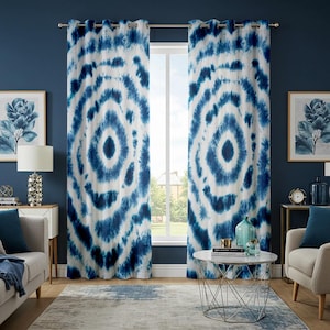Op de afbeelding: Twee blauw-witte gordijnen met een cirkelvormig tie-dye patroon hangen voor een raam. De gordijnen hebben een ontwerp met een donkerblauw centrum en lichtere blauwe ringen. De kamer heeft een blauwe muur, een grijs tapijt en een witte tafel.