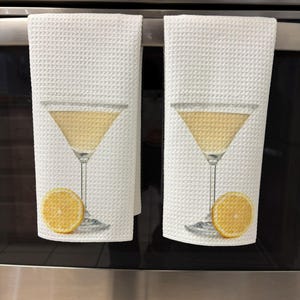 May include: Two white kitchen towels with a yellow lemon slice and a martini glass printed on them. The towels are hanging on a stainless steel oven.
