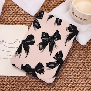 May include: A light pink tablet case featuring a pattern of black bows. The bows are stylized with brushstroke details. A small card with the word "EMBRACE" is visible. A white mug and napkin are in the background.