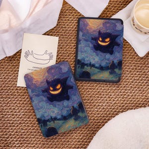 May include: Two tablet cases and a small card rest on a woven surface. The tablet cases feature a dark purple cartoon ghost with a glowing orange grin against a swirling blue and yellow background. The card has a line drawing of two hands and the text "EMBRACE Can you give".