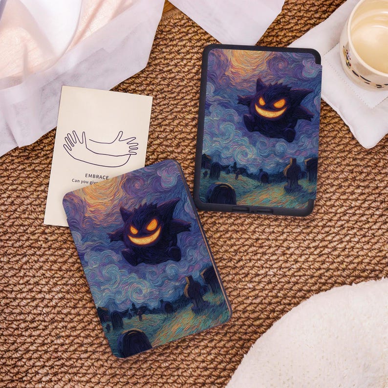 May include: Two tablet cases and a small card rest on a woven surface. The tablet cases feature a dark purple cartoon ghost with a glowing orange grin against a swirling blue and yellow background. The card has a line drawing of two hands and the text "EMBRACE Can you give".