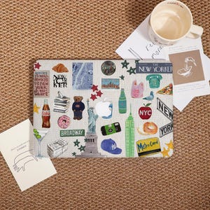 May include: A laptop sleeve featuring a collage of New York City-themed stickers. The design includes images of the Statue of Liberty, the Empire State Building, and the text "The New Yorker". Other stickers include a bear, a donut, and a MetroCard.