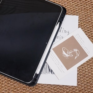 May include: A black tablet with a white stylus is placed on top of two pieces of paper. One piece of paper has a brown square with a white illustration of hands holding a bowl. The other piece of paper has the word "ding" and "min" printed on it.