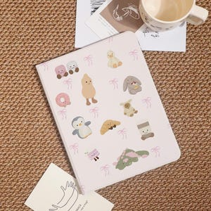 May include: A light pink notebook with various cartoon illustrations, including a penguin, duck, and bunny. The notebook is decorated with small pink bows. A white mug and a card are in the background.