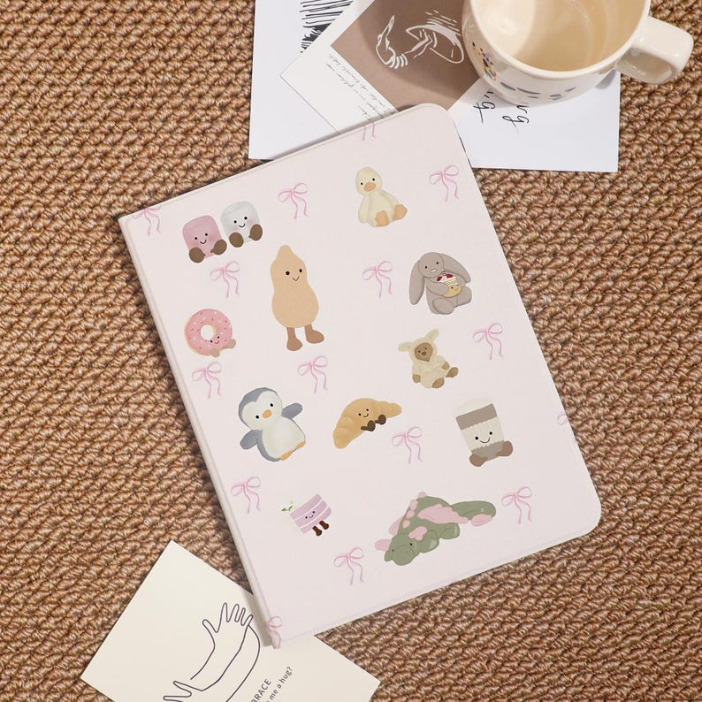 May include: A light pink notebook with various cartoon illustrations, including a penguin, duck, and bunny. The notebook is decorated with small pink bows. A white mug and a card are in the background.