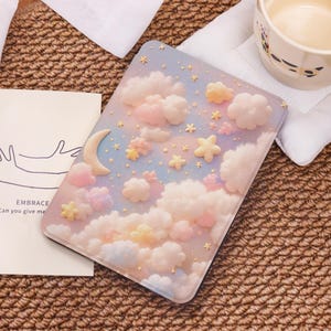 May include: A pastel pink and blue e-reader cover with a whimsical design of clouds, stars, and a crescent moon. The clouds are 3D and have a fluffy texture.