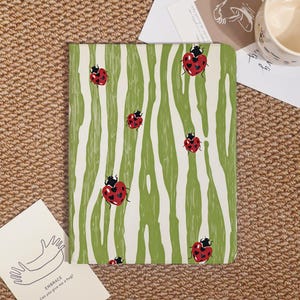 May include: A rectangular tablet case featuring a pattern of red ladybugs with black spots on a background of vertical, wavy light green and white stripes. The ladybugs are shaped like hearts, and the case has rounded corners.
