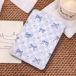 May include: A light blue and white checkered tablet case with a pattern of blue bows and hearts. The case is rectangular and has a soft, padded appearance. The case is on a woven surface.