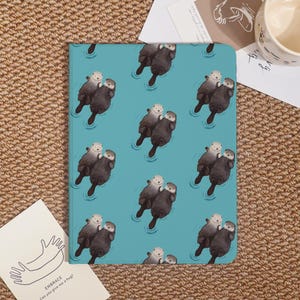 May include: A notebook with a teal background featuring a repeating pattern of otters floating in water. The otters are depicted in a cartoon style, with one otter holding another. The notebook is rectangular and appears to be closed.