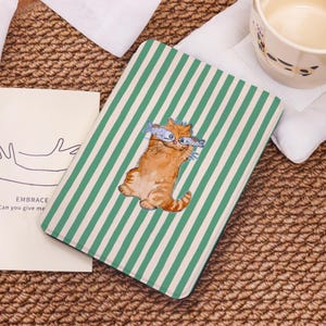 May include: A rectangular notebook with a green and white striped pattern. The cover features an orange cat illustration holding two fish. The notebook is on a woven surface with a white cloth and a ceramic mug.