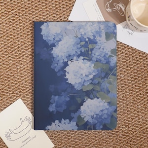 May include: A notebook with a blue floral design featuring white and blue hydrangea flowers. The notebook has a dark blue background and a soft, watercolour-like aesthetic. A card with the word "EMBRACE" and the phrase "Can you give me a hug?" is in the lower left corner.