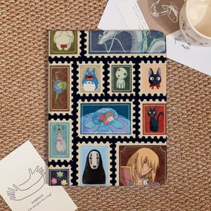 May include: A patterned notebook featuring various anime characters in a stamp-like design. The notebook has a black border and a beige background. The characters include a blue creature, a black cat, and a girl in a bubble.