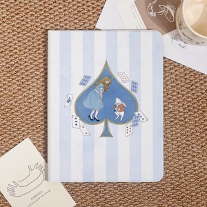 May include: A notebook with a blue and white striped background. The cover features an illustration of Alice in Wonderland inside a spade shape, with playing cards scattered around. The notebook is a whimsical item.