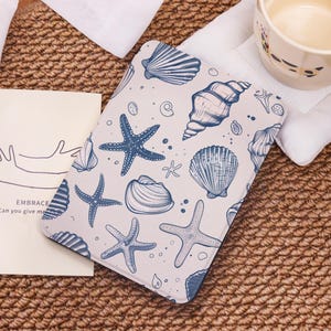 Könnte beinhalten: Ein rechteckiger Gegenstand mit hellem Hintergrund und einem Muster aus blauen Muscheln und Seesternen. Das Design zeigt verschiedene Muschelformen und Seesterne. Der Gegenstand liegt auf einer gewebten Oberfläche.