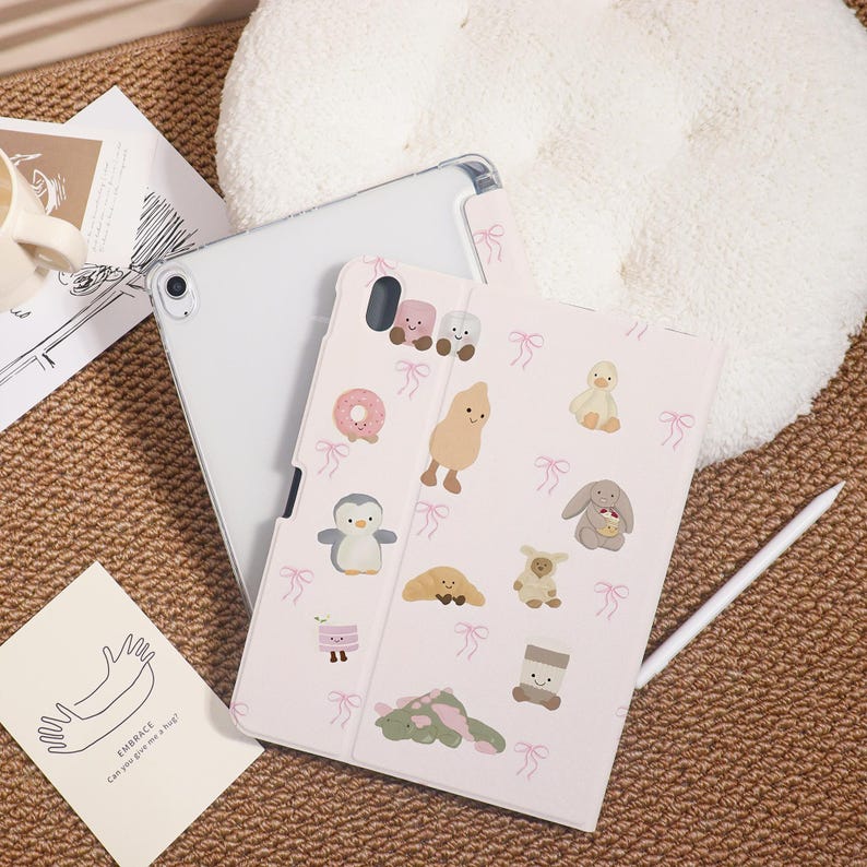 May include: A light pink tablet case with a pattern of cartoon characters and pink bows. The case is open, revealing a white tablet. A white stylus rests on the case. A card with the word "EMBRACE" is visible.