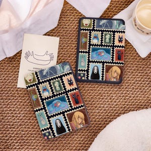 May include: Two black and blue phone cases with a stamp design featuring anime characters. The cases are on a woven surface with a card that says "EMBRACE" and a drawing of hands. A cup of liquid is in the background.