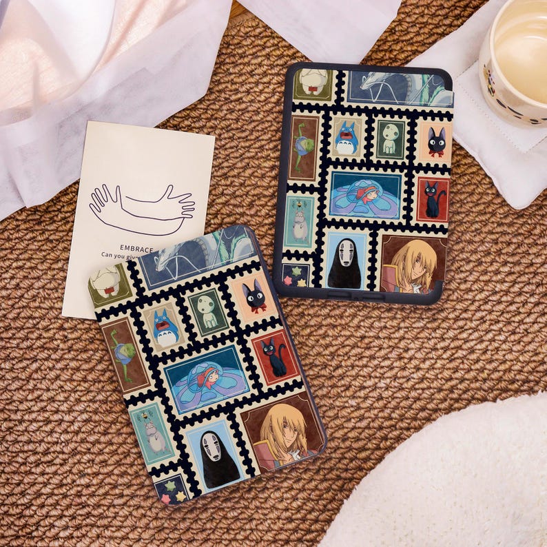 May include: Two black and blue phone cases with a stamp design featuring anime characters. The cases are on a woven surface with a card that says "EMBRACE" and a drawing of hands. A cup of liquid is in the background.