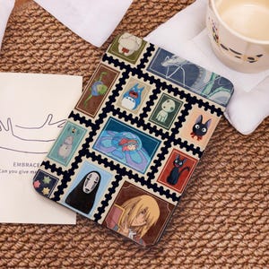 May include: A black tablet case with a collage of colourful anime-style illustrations resembling postage stamps. The case has a black border and a beige background. The text "EMBRACE Can you give me" is visible on a card.