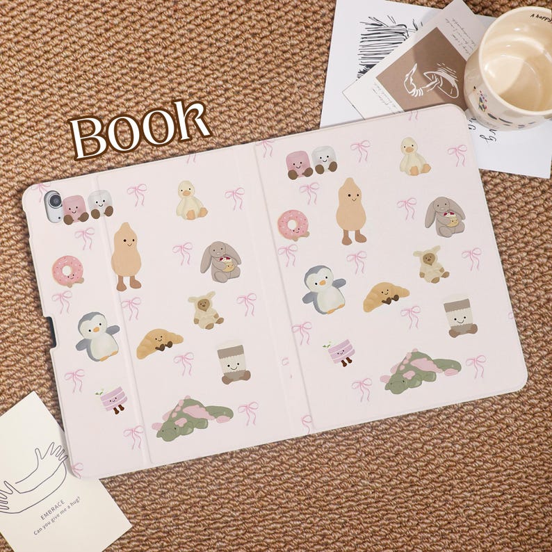 May include: A light pink tablet case with a repeating pattern of cute cartoon characters, including ducks, bunnies, and penguins. The word "BOOK" is printed in large letters at the top. A mug and a card are in the background.
