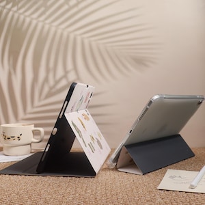 May include: Two tablet cases are displayed, one black and one clear. The black case is open, revealing a patterned interior with botanical illustrations. The clear case is also open, showcasing the tablet's screen. A mug and a pen are also visible.