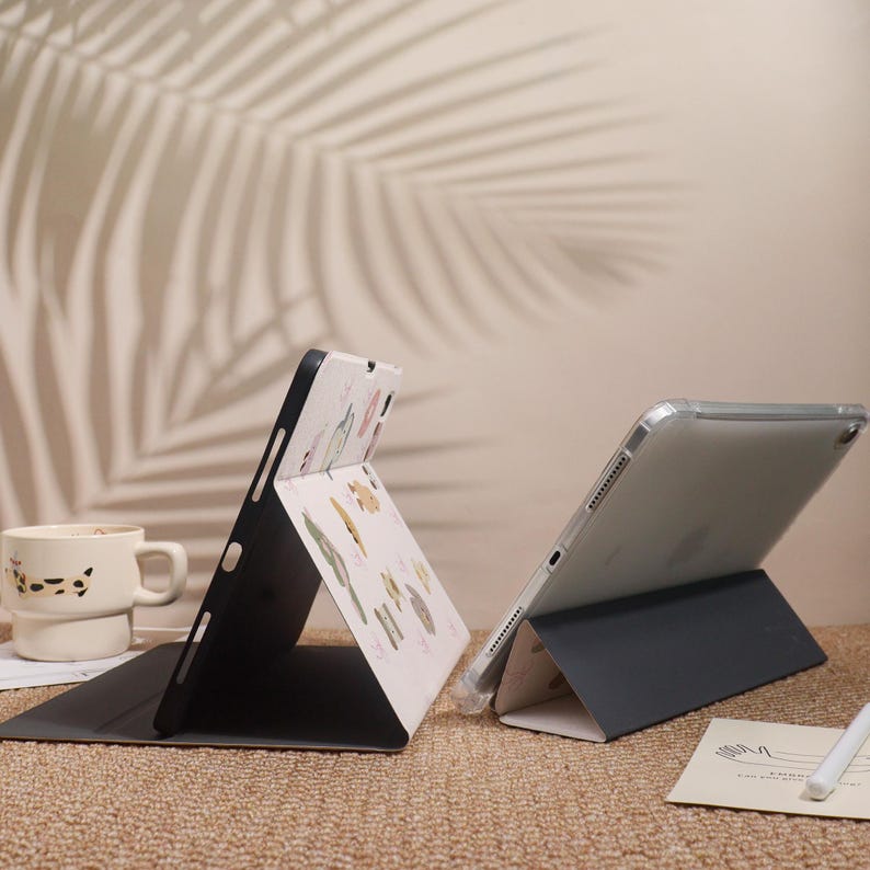 May include: Two tablet cases are displayed, one black and one clear. The black case is open, revealing a patterned interior with botanical illustrations. The clear case is also open, showcasing the tablet's screen. A mug and a pen are also visible.