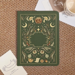 May include: A dark green hardcover journal featuring a gold-toned illustration of a central blank frame surrounded by botanical elements, mushrooms, a cauldron, crescent moons, a mortar and pestle, a scroll, and acorns in a symmetrical arrangement.