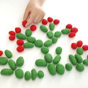 Op de afbeelding: Een kinderhand reikt naar een rode vilten bal tussen een verzameling groene en rode vilten vormen. De vormen, een mix van rond en ovaal, zijn gerangschikt op een wit oppervlak. Viltstukken voor een knutsel- of educatieve activiteit.