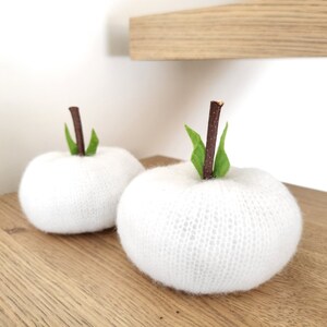 Peut inclure: Deux décorations en forme de pomme, tricotées en blanc, avec des tiges marron et des feuilles vertes. Les pommes sont posées sur une surface en bois, avec un fond blanc. La texture tricotée est chaleureuse.
