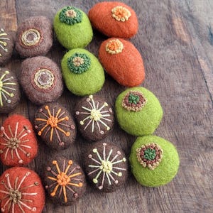 May include: An assortment of colorful, oval-shaped felted stones. The stones are in shades of brown, orange, and green, with embroidered floral designs in various colors. The stones are arranged on a wooden surface.