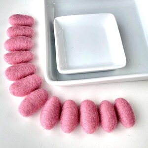 Puede incluir: Cuentas de lana afieltrada rosa dispuestas cerca de dos platos de cerámica blanca. Las cuentas de forma ovalada tienen una superficie texturizada. Los platos son cuadrados y rectangulares, creando una estética minimalista.