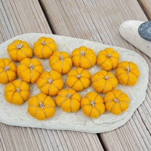 May include: A collection of small, bright orange felt pumpkins with silver stems arranged on a light gray stone. The pumpkins are clustered together, suggesting a harvest or autumn theme. A white and gray stone are in the background, set against a wooden surface.