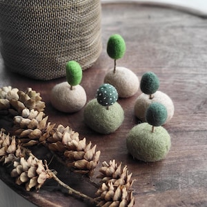 Può includere: Ornamenti per alberi in feltro fatti a mano in varie tonalità di verde e beige, disposti su un vassoio di legno. Gli ornamenti hanno piccole cime rotonde a forma di albero. Sono presenti anche pigne e un cestino intrecciato pieno di piccoli bastoncini di legno.