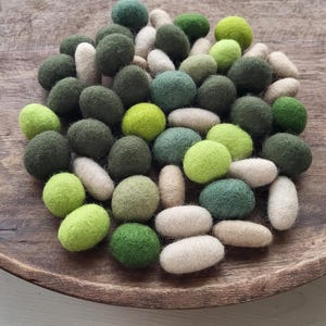 Op de afbeelding: Een verzameling vilten wollen ballen en langwerpige vormen in verschillende tinten groen en gebroken wit, gerangschikt in een rustieke houten kom. De kleuren variëren van licht limoengroen tot diep bosgroen.