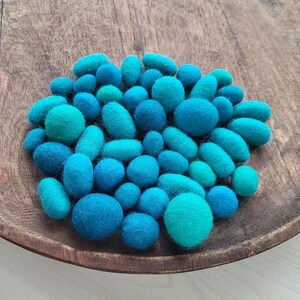Può includere: Una ciotola di legno piena di forme di lana infeltrita blu e turchese. Le forme includono sfere e ovali in varie tonalità di blu. La ciotola ha una finitura a venatura naturale. Un'esposizione vibrante.
