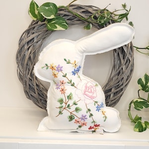 Könnte beinhalten: Ein weißes, hasenförmiges Kissen mit gestickten Blumen in Rosa, Gelb, Blau und Orange. Es steht vor einem grauen Weidenkranz und Grünzeug, was ein Frühlings- oder Osterthema ergibt. Ein dekoratives Element.
