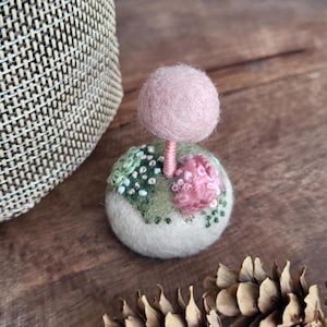 Könnte beinhalten: Eine handgefertigte Filz-Pilz-Skulptur mit rosa Kappe und Stiel, auf einem runden Sockel mit gestickten Blumen- und Blattdetails. Die Skulptur steht auf einer Holzoberfläche, mit einem geflochtenen Korb und einem Tannenzapfen im Hintergrund.