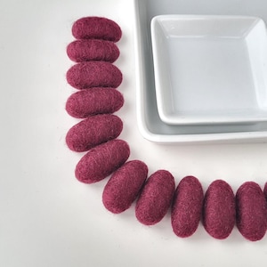 Puede incluir: Una colección de cuentas de fieltro de color rojo oscuro, de forma ovalada, dispuestas en una línea curva. Dos platos de cerámica blanca apilados en el fondo. Las cuentas tienen un aspecto texturizado, sobre un fondo blanco.