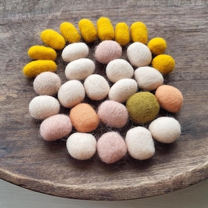 May include: A selection of colourful felt balls and oblong shapes in a wooden bowl. The felt pieces are in shades of yellow, white, pink, orange, and green. The arrangement is on a dark wooden surface.