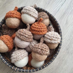 Könnte beinhalten: Ein Korb voller handgefertigter Eichel-Ornamente. Die Eicheln haben weiße oder beige Körper und gehäkelte Kappen in verschiedenen Braun- und Orangetönen. Der Korb steht auf einer hellen Holzoberfläche.