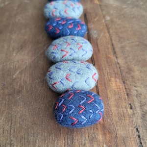 May include: A vertical arrangement of six oval-shaped felted stones. The stones alternate between shades of blue and gray, each adorned with a stitched chevron pattern in contrasting colors of red and purple. The stones are displayed on a wooden surface.