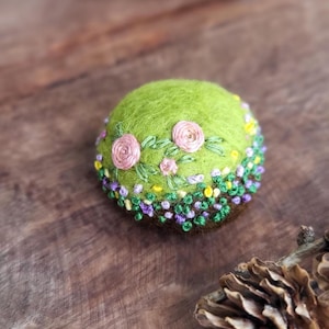 May include: A round, green felted object with embroidered floral details. Two pink roses are centered on the top, surrounded by green leaves and small yellow and purple flowers. The base is brown, and the object is set on a wooden surface.