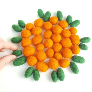 Puede incluir: Vista cenital de una composición de bolas de fieltro naranjas y verdes sobre fondo blanco. Las bolas naranjas se agrupan en el centro, rodeadas de formas ovaladas verdes. Se ve una mano sosteniendo una de las bolas de fieltro verdes.