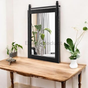 May include: A black framed mirror with a sliding barn door style design. The mirror is hanging on a white wall above a wooden table.