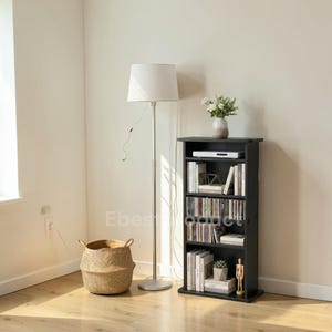 May include: A black bookshelf with multiple shelves, filled with books and CDs. A white lamp with a cylindrical shade stands next to the shelf. A woven basket sits on the floor, and a vase of flowers is on top of the shelf.