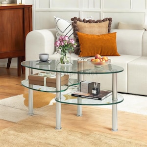 May include: A glass coffee table with two tiers. The table has a unique, asymmetrical shape and is supported by four silver metal legs. The top tier is decorated with a vase of pink flowers, a cup and saucer, and a bowl of fruit. The bottom tier has a box with the word "Chanel" on it, a book, and a set of reed diffusers.