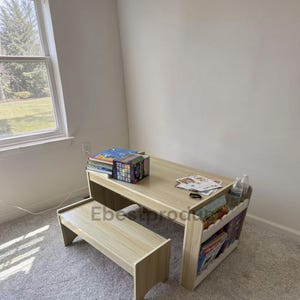 May include: A light wood-grain children's activity table and bench set featuring a flat tabletop surface, a side storage compartment for books, and a matching bench. The table holds a stack of books, a box, and papers, with water bottles on the side.